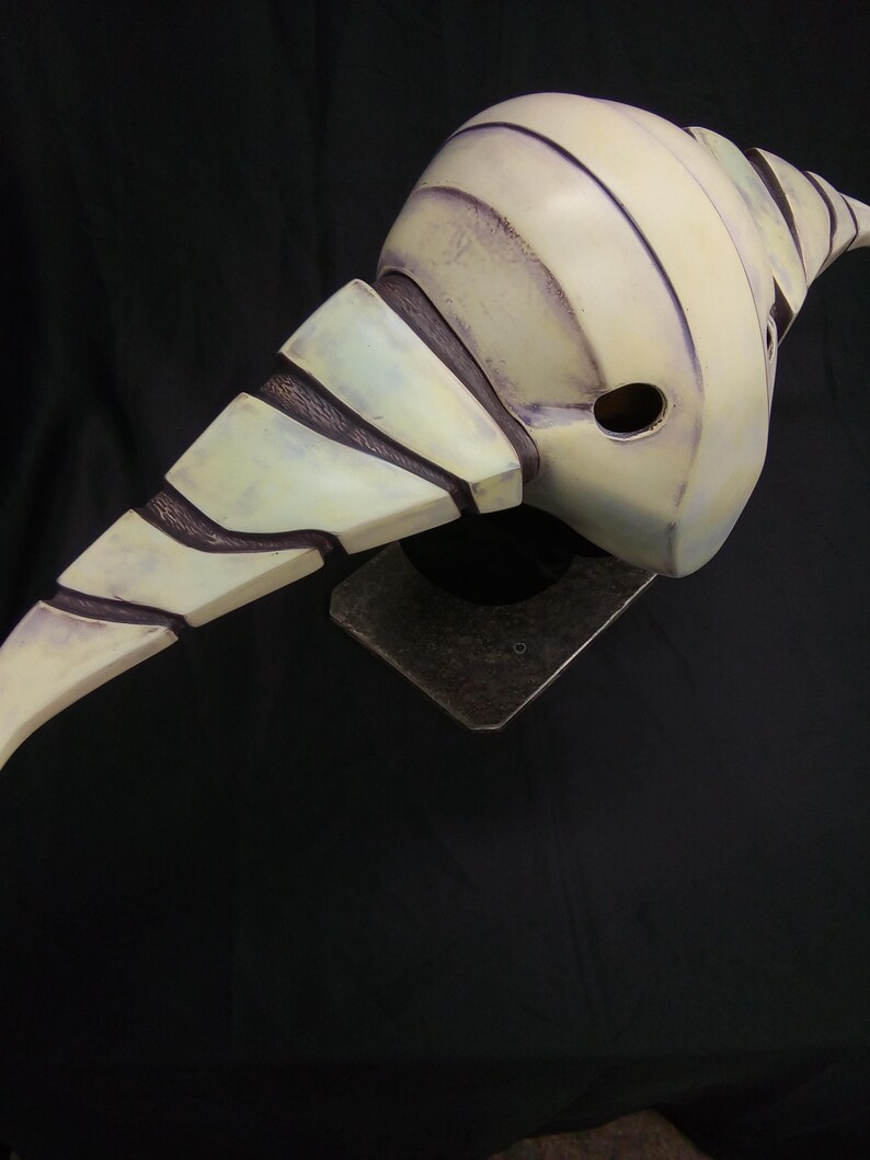 May include: A white and purple mask with a textured surface and a pointed, curved design. The mask has a small hole on the side.