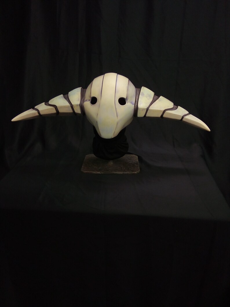 May include: A white and black mask with horns, resembling a creature from a fantasy world. The mask has a textured surface and two eye holes.
