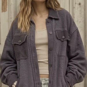 Può includere: Una giacca a camicia in pile grigio antracite scuro con chiusura a bottoni, colletto, maniche lunghe con polsini a coste, due tasche sul petto con bottoni e due tasche laterali, indossata sopra una canotta a righe beige e jeans chiari.