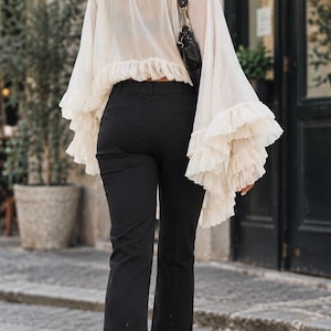 May include: A cream-colored long-sleeve blouse with a high neck and tiered ruffled bell sleeves paired with black cropped trousers featuring a small vertical slit at the hem and a black shoulder bag with a thin strap and silver hardware.