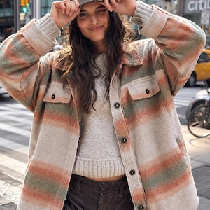 May include: A long-sleeved button-down shirt jacket featuring a horizontal striped pattern in muted shades of orange, green, and cream. The garment has a fold-over collar, two chest pockets with button closures, and ribbed cuffs on the sleeves.