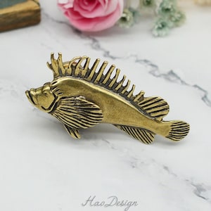 Könnte beinhalten: Ein goldfarbener Fisch-Anhänger aus Metall mit detaillierten Flossen und Schuppen. Der Fisch blickt nach links und hat eine stachelige Rückenflosse. Der Anhänger liegt auf einer weißen Marmorfläche.