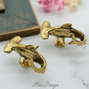 May include: Two brass-colored hammerhead shark cabinet knobs. The knobs are detailed with fins and a tail, and have a screw post on the back. The knobs are on a white surface.
