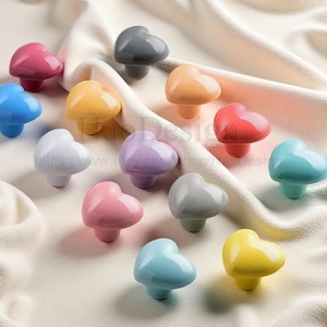 Pode incluir: Uma coleção de puxadores de armário em forma de coração em várias cores, incluindo rosa, azul, amarelo e cinza. Os puxadores estão dispostos sobre um tecido macio de cor creme, mostrando seu acabamento brilhante e design decorativo.
