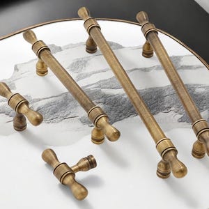 May include: A selection of antique brass cabinet handles and knobs. The image shows long bar pulls and smaller T-shaped knobs, displayed on a white and grey marble-effect surface. The brass hardware has an aged finish, highlighting the detailed design.