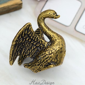 May include: A gold-toned swan figurine with detailed wings and a textured surface. The swan is positioned with its head turned to the side and its wings spread out.
