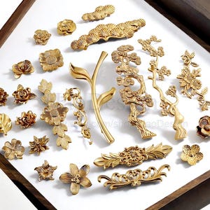 以下が含まれることがあります： 金色の装飾用金具のコレクション。キャビネットの引き手、ノブ、装飾的なアクセントが含まれています。 金具は、花柄、葉柄、枝柄のデザインが特徴です。 作品は木製のトレイ内の白い表面に配置されています。