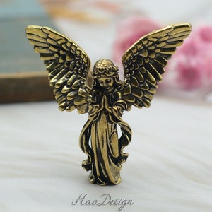 May include: A small, gold-colored metal angel figurine with outstretched wings. The angel is wearing a long robe and has its hands clasped in prayer. The figurine is standing on a white surface.