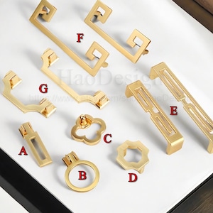 May include: A selection of gold-coloured cabinet hardware, including handles and knobs. The hardware features various geometric designs, such as rectangular, square, and clover shapes. The image is taken from a top-down perspective.