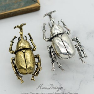 Peut inclure: Deux figurines de scarabées en métal, une dorée et une argentée, sont présentées sur une surface blanche. Les scarabées ont des pattes, des antennes et une protubérance en forme de corne détaillées. L'image comprend le texte "Hao Design" et une adresse de site web.