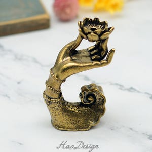 May include: A gold-toned metal incense burner shaped like a hand holding a lotus flower. The hand is adorned with intricate details and the lotus flower has a small opening in the centre for incense.