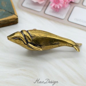Puede incluir: Una manija de puerta o tirador en forma de ballena de color dorado. La ballena está detallada con una superficie texturizada y una boca ligeramente abierta.