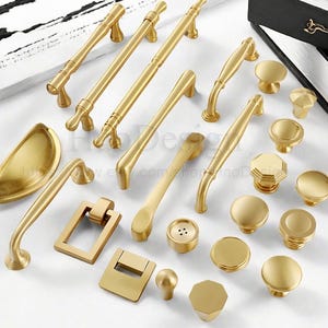 May include: A collection of gold-colored cabinet hardware, including handles, knobs, and pulls. The hardware features various designs, from simple to ornate, with a matte finish. The assortment includes different shapes and sizes for diverse furniture applications.