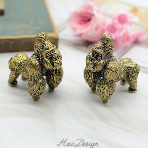 May include: Two small gold-colored gorilla figurines. The figurines are detailed and have a textured surface. They are standing on a white surface.