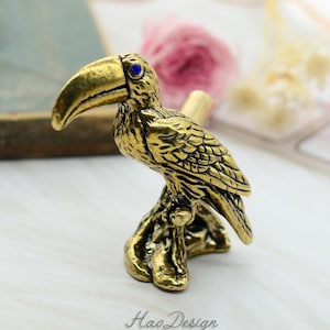 Könnte beinhalten: Eine goldene Tukan-Figur mit einem blauen Auge, auf einem Ast sitzend. Der Schnabel des Tukans ist geöffnet und die Figur ist mit filigranen Federn verziert.