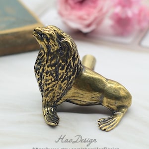 Puede incluir: Una figurita de león marino de metal dorado con una superficie texturizada detallada. La figurita está en posición sentada con la cabeza inclinada hacia arriba. Es una pieza decorativa que podría usarse como tirador de cajón o pomo de armario.