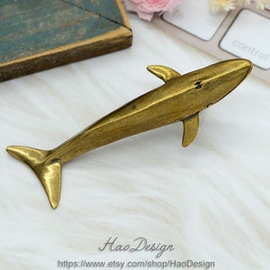 Könnte beinhalten: Eine goldfarbene Metallfigur eines Wals. Der Wal blickt nach rechts im Bild und steht auf einer weißen Marmorfläche.