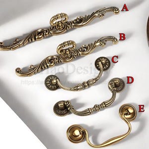 Puede incluir: Una variedad de tiradores y manijas de cajón de latón antiguo. La colección incluye tiradores curvos y ornamentados con detalles decorativos y perillas redondas de inspiración floral. El hardware se muestra sobre una superficie blanca.