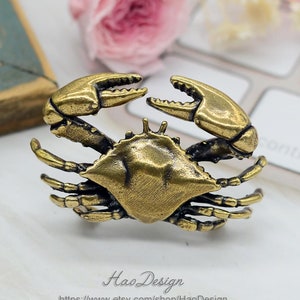 Könnte beinhalten: Eine goldfarbene Metallfigur eines Krabben mit detaillierten Klauen und Beinen. Der Krebs ist mit erhobenen Klauen positioniert, als ob er zum Zupacken bereit wäre.
