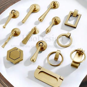 Peut inclure: Une collection de quincaillerie de meubles dorée, comprenant des poignées, des tirettes et des boutons. La quincaillerie présente divers modèles, tels que des anneaux de tirage, des barres de tirage et des poignées à levier. Les pièces sont disposées sur une surface blanche.