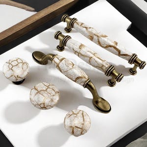 Puede incluir: Un conjunto de herrajes para armarios, que incluye tres tiradores blancos y dorados con diseño de mármol y tres pomos redondos a juego. Los tiradores tienen un diseño curvo con detalles en color latón, y los pomos son esféricos.