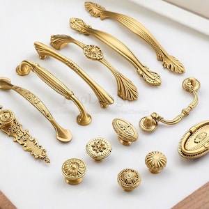 Può includere: Una collezione di ferramenta dorata per mobili, tra cui maniglie e pomelli. La ferramenta presenta dettagliati disegni floreali e geometrici, con varie forme e dimensioni, esposti su una superficie bianca.