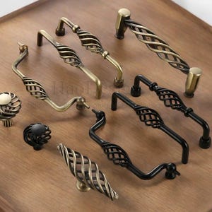 以下が含まれることがあります： アンティーク真鍮と黒仕上げの、装飾的なキャビネットノブとプル。ケージのようなデザインとスパイラルデザインが特徴です。木製の表面に展示されています。画像に寸法は含まれていません。