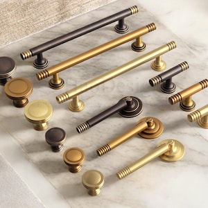 May include: A selection of cabinet handles and knobs in gold, bronze, and black finishes. The image shows a variety of designs, including bar pulls and round knobs, displayed on a marble surface.