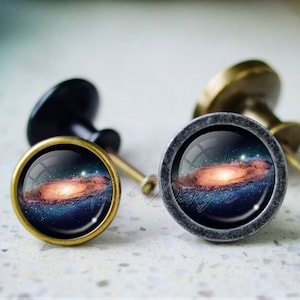 以下が含まれることがあります： 銀河の画像が中央に描かれたキャビネットノブが2つ。ノブは金属製で、ビンテージな外観です。1つのノブはアンティークブラス、もう1つはアンティークシルバーです。