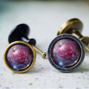 May include: Two cabinet knobs with a galaxy design. The knobs are round and have a silver finish. The galaxy design is a nebula with pink, purple, and blue colors.