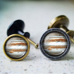 May include: Two cabinet knobs with a round, brass base and a glass top featuring a detailed image of Jupiter. The knob on the left has a brass finish, while the knob on the right has a dark gray finish.