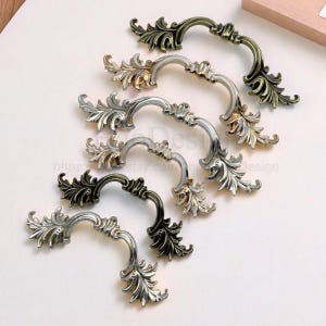May include: An assortment of ornate cabinet pulls in various metallic finishes, including antique brass, silver, and gold. Each pull features a curved handle and detailed leaf-like embellishments, showcasing a vintage aesthetic. The hardware is arranged on a white surface.