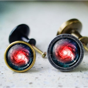 May include: Two cabinet knobs featuring a galaxy design. The knobs are made of metal and have a dark grey finish. The galaxy design features a swirling red and blue nebula.