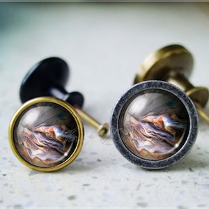 Puede incluir: Dos pomos de armario redondos con una base metálica y una parte superior de vidrio que presenta un diseño abstracto en espiral en tonos marrones, naranjas y morados.