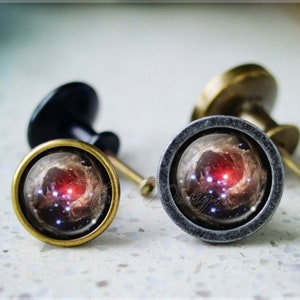 Puede incluir: Dos pomos decorativos para armarios con un diseño de galaxia. Los pomos son de metal y tienen forma redonda. El diseño de galaxia presenta una nebulosa giratoria con colores rojo, rosa y azul.