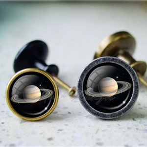 May include: Two cabinet knobs with a black background and a gold ring around the edge. The knobs feature a depiction of Saturn with its rings.