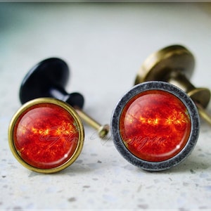 Puede incluir: Dos pomos de armario con una imagen de sol roja y naranja. Los pomos son de metal con acabado en latón. Un pomo tiene una base negra y el otro una base plateada.