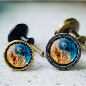 Puede incluir: Dos pomos de armario con un diseño de yin yang. Los pomos son de metal y tienen un acabado negro y plateado. El diseño de yin yang es un remolino azul y naranja.