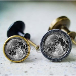 Puede incluir: Dos pomos de armario con una imagen de la luna. El pomo de la izquierda tiene un acabado en latón y el pomo de la derecha tiene un acabado en plata.