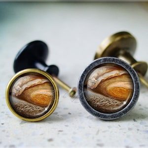 Puede incluir: Dos pomos de armario con acabado metálico. Los pomos presentan un diseño redondo y elevado con una imagen impresa de las nubes de Júpiter en tonos marrones, naranjas y blancos.