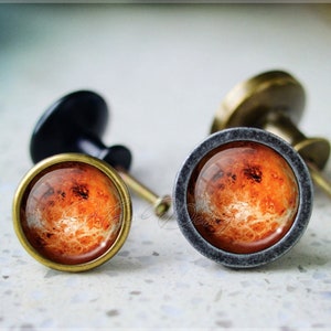 Puede incluir: Dos pomos de armario con acabado metálico. Los pomos presentan una superficie redonda, similar al vidrio, con una imagen detallada de un planeta. El planeta es de color rojo-naranja con nubes blancas.