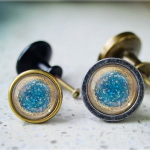 Puede incluir: Dos pomos decorativos para armarios con incrustaciones de resina azul y dorada. Los pomos son redondos y tienen una base metálica.