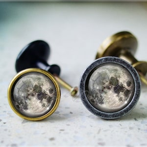May include: Two cabinet knobs featuring a moon image. The knobs are made of metal with a black and grey finish. The moon image is in shades of grey and white.
