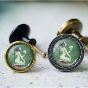 May include: Two antique-style cabinet knobs with a round, silver-toned base and a gold-toned top. Each knob features a circular, glass-like insert with a green background and a fairy illustration holding a white flower.