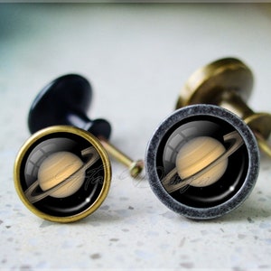Peut inclure: Deux boutons de porte avec une finition dorée et une base noire. Les boutons présentent un insert en verre rond avec une image de Saturne et ses anneaux.