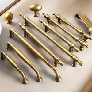 May include: A collection of antique brass cabinet hardware, including knobs and various sizes of pull handles. The hardware has a warm, aged finish and a classic design. The arrangement showcases different styles for kitchen or furniture projects.