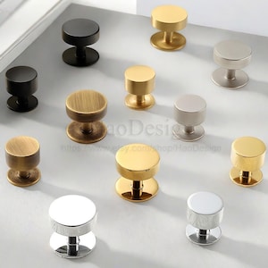May include: A collection of cabinet knobs in various metallic finishes, including gold, silver, and black. The knobs have a cylindrical design with a flat top and base, suitable for furniture hardware.
