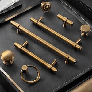 Può includere: Una collezione di ferramenta per mobili in ottone antico, tra cui pomelli, maniglie e un tirante ad anello. La ferramenta ha una finitura strutturata e una calda tonalità dorata. I pezzi sono disposti su una superficie grigio scuro.