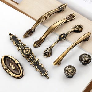 Può includere: Un assortimento di ferramenta per mobili in ottone antico, tra cui maniglie e pomelli. Le maniglie variano per dimensioni e design, con dettagli ornati e una finitura calda e invecchiata. I pomelli presentano motivi floreali.