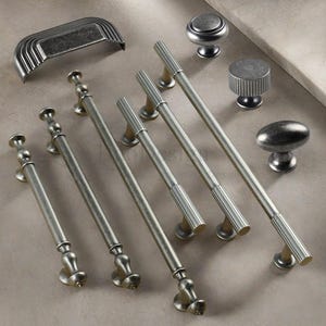 Puede incluir: Una variedad de herrajes para gabinetes de estilo antiguo. La imagen muestra varios tiradores largos y acanalados, perillas decorativas y un tirador de taza, todos en un acabado plateado apagado. El hardware está dispuesto sobre una superficie neutra.